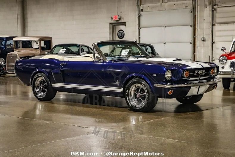 A classic blue and white convertible Mustang, a high-performance gem, sits in a garage showroom surrounded by other vintage muscle cars.