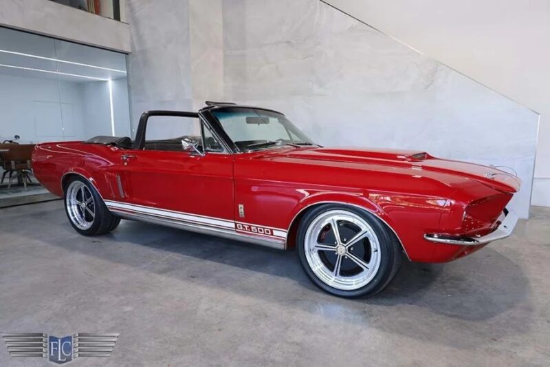 A red vintage convertible with GT500 markings, a black soft top, and high-performance features is parked in a glossy-floored garage with white walls. Perfect for collectors of classic muscle cars, it might just be your dream ride for sale.