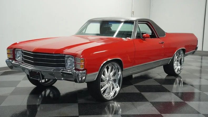 A red vintage car with a black roof and large chrome wheels is parked indoors on a checkered floor, embodying the allure of classic muscle cars. This high-performance beauty is now for sale to collectors who appreciate timeless elegance with a powerful edge.