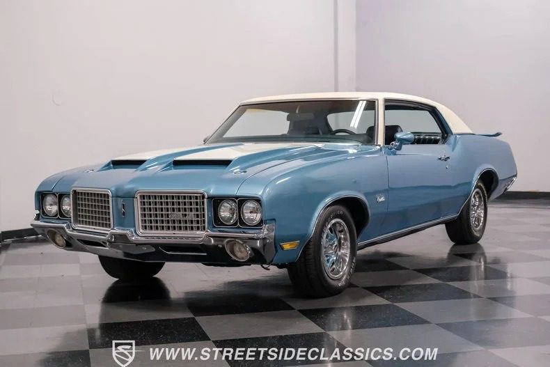 A blue vintage car with a white roof, showcasing a classic design and chrome details, sits parked indoors on a checkered floor—a true gem among classic muscle cars.