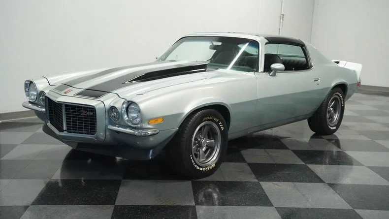 A silver and gray high-performance muscle car with a black stripe on the hood, parked indoors on a checkered floor.