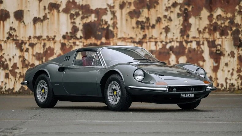 A vintage gray Ferrari Dino is parked in front of a rusted metal wall.