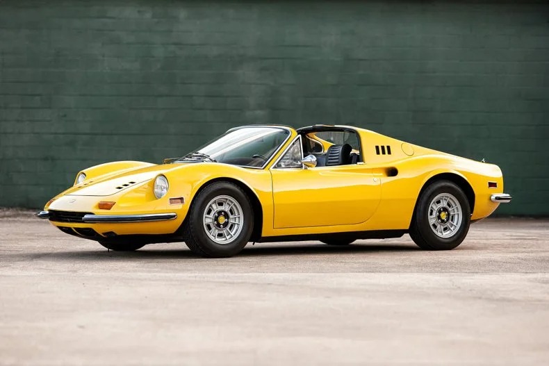 A yellow vintage Ferrari Dino sports car is elegantly parked in front of a green wall, echoing a rich history of automotive excellence.