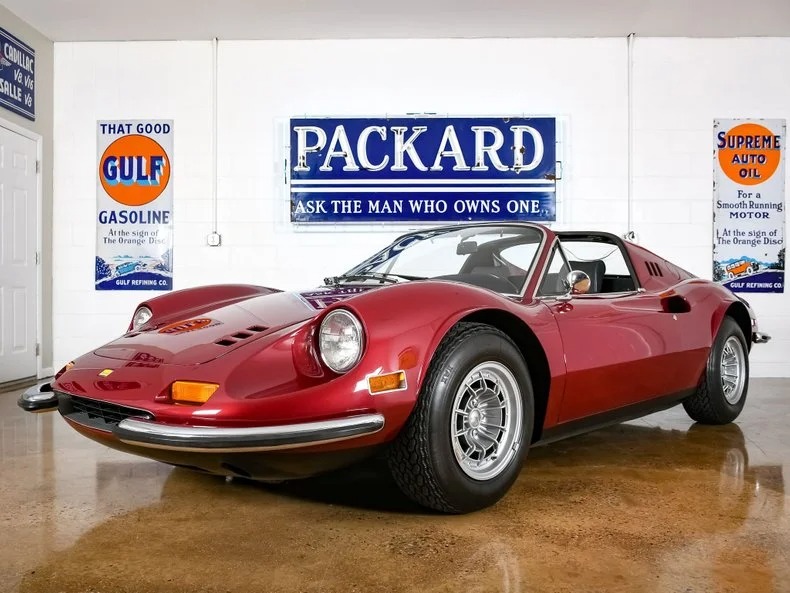 A red vintage sports car, resembling a classic Ferrari Dino, is parked indoors in front of a white wall adorned with retro signs like "Packard" and "Gulf," painting a picture steeped in automotive history.