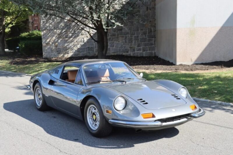 A classic silver Ferrari Dino sports car is parked on a street, with a stone wall and trees in the background.