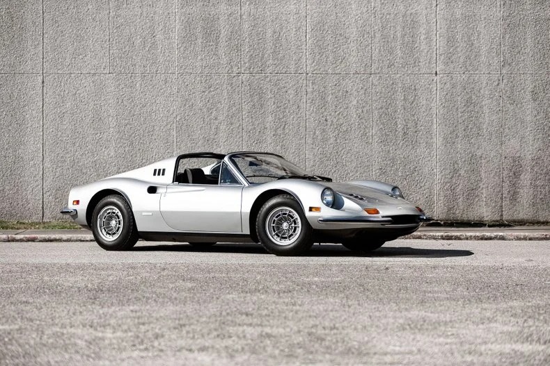 A classic silver convertible Ferrari Dino sports car is parked in front of a gray textured wall, capturing the essence of timeless elegance and sophistication.