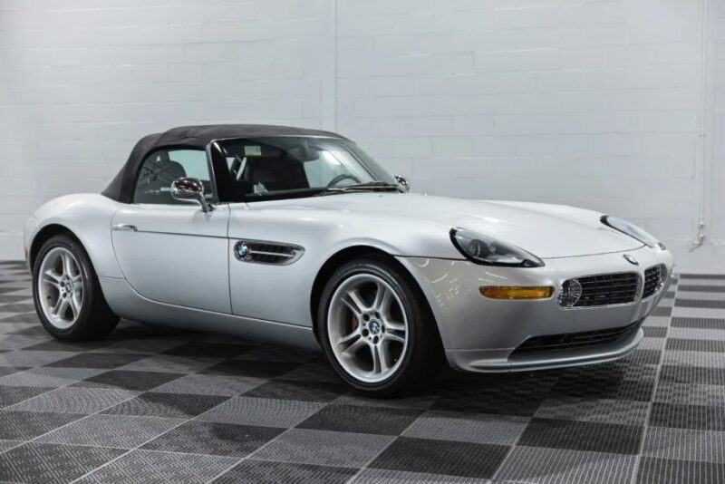 A silver BMW Z8 with a black convertible top is parked indoors on a checkered floor, offering an open-air modern classic experience.