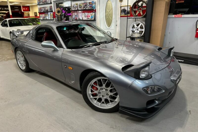 A gray sports car with pop-up headlights sits in the garage, surrounded by various automotive memorabilia and car parts. Capturing that new nostalgia of 2000s cars, it's a classic find that's now up for sale.