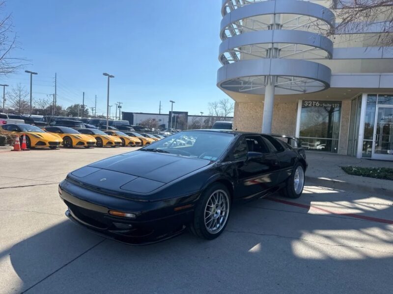 A sleek black sports car, reminiscent of the best 2000s cars for sale, is parked in front of a modern building. In the sunny backdrop with clear skies, several yellow cars evoke a sense of New Nostalgia.