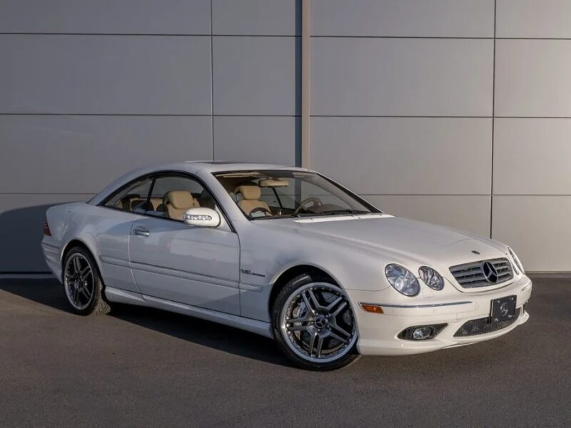 Coupe Mercedes-Benz putih mewah diparkir dengan elegan di permukaan beton, dibingkai oleh dinding ubin abu-abu yang ramping.