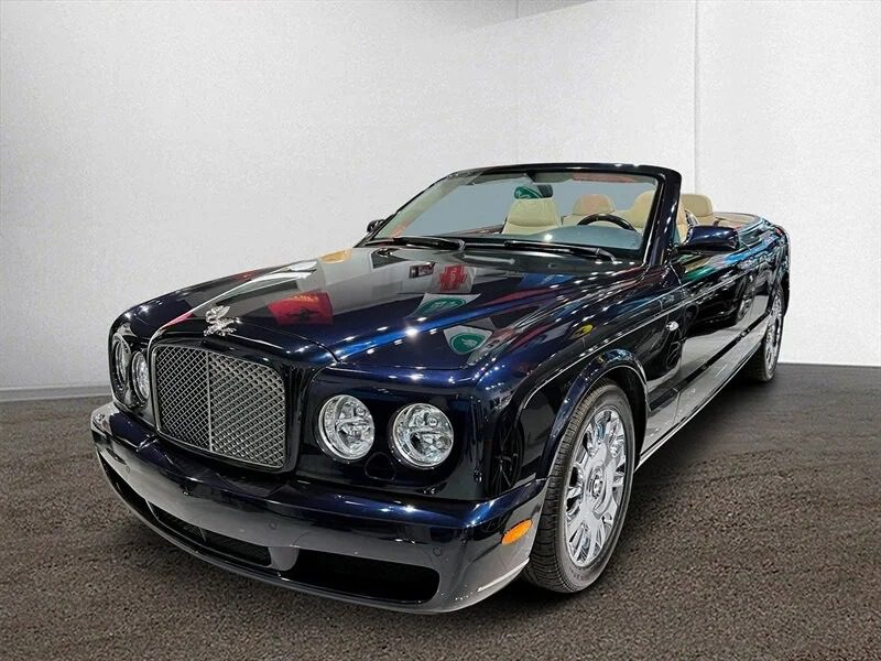 A dark-colored luxury convertible, reminiscent of the best 2000s cars, is parked indoors on a carpeted floor. Its top is down, revealing a light tan interior that evokes a sense of nostalgia for classic drives.