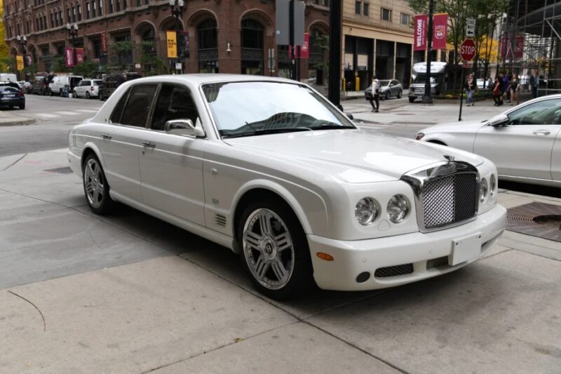 A white luxury sedan, reminiscent of the best cars from the 2000s, is parked on a city street, surrounded by elegant buildings and lush greenery.