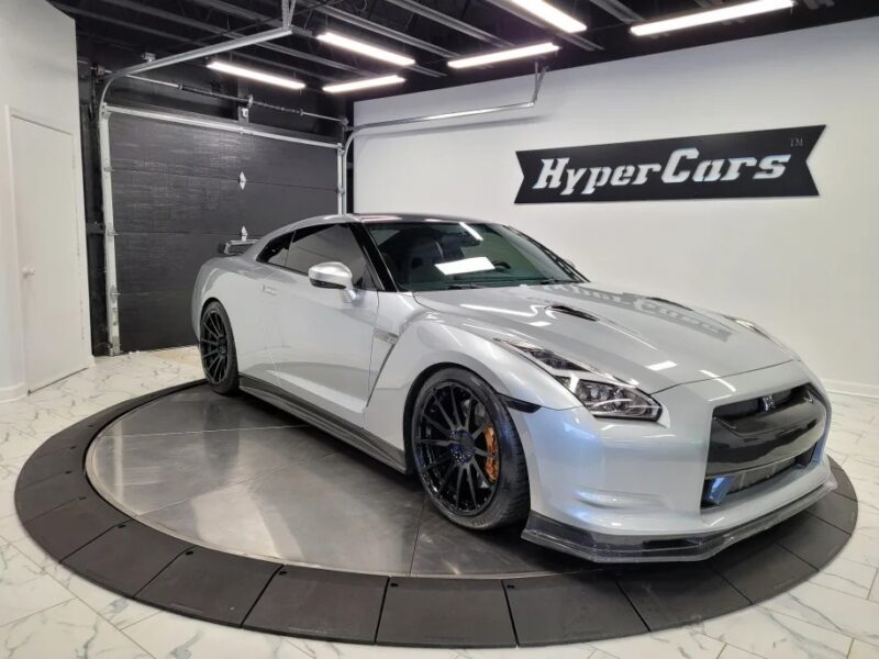 A silver sports car from the 2000s graces the rotating platform in an indoor showroom, exuding nostalgia. With "HyperCars" boldly displayed on the wall, it's unmistakably for sale to those who cherish past eras of automotive excellence.