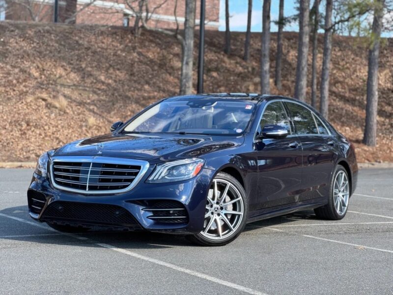 Sedan Mercedes-Benz biru tua, yang melambangkan keanggunan, diparkir di permukaan beraspal dengan pohon-pohon dan bangunan bata di latar belakang.