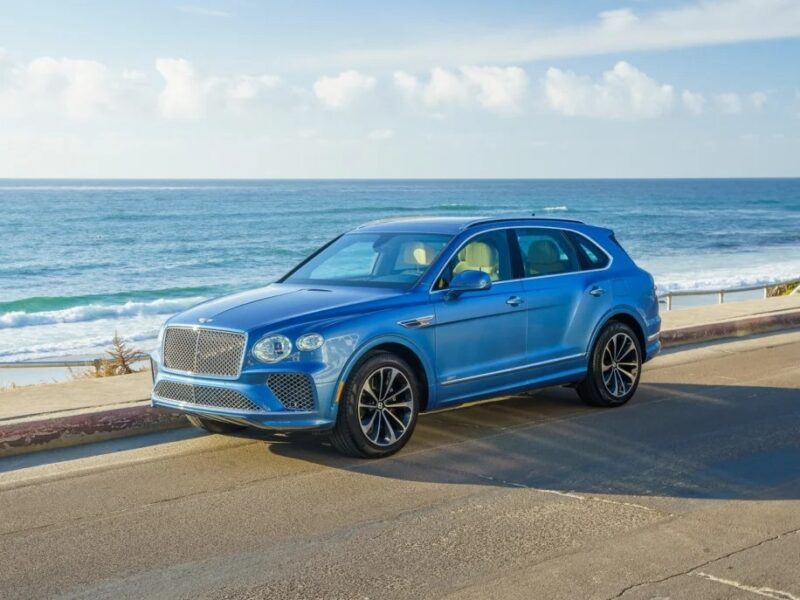 A blue Bentley Bentayga is gracefully parked on a coastal road, with the vast ocean and serene sky painting an exquisite backdrop for this ultra-luxe SUV.