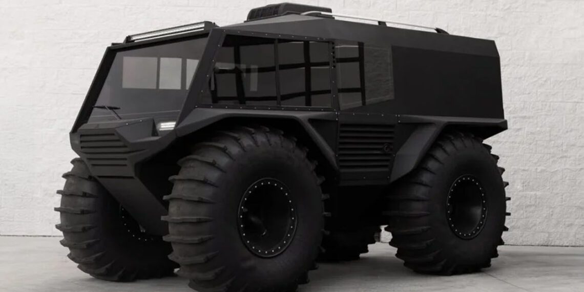 A futuristic black all-terrain vehicle with large tires is parked on a concrete surface against a white brick wall.