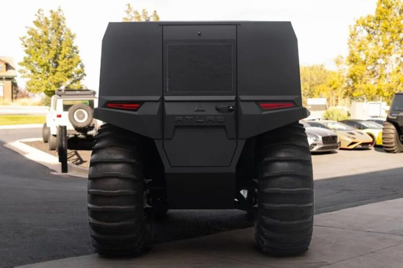 Back view of a large, matte black off-road vehicle with oversized tires and minimal branding, parked outside on a sunny day.