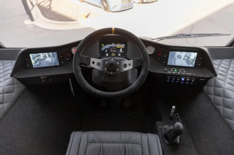 Interior view of a vehicle's cockpit showing a steering wheel, digital dashboard, side cameras, and gear lever.