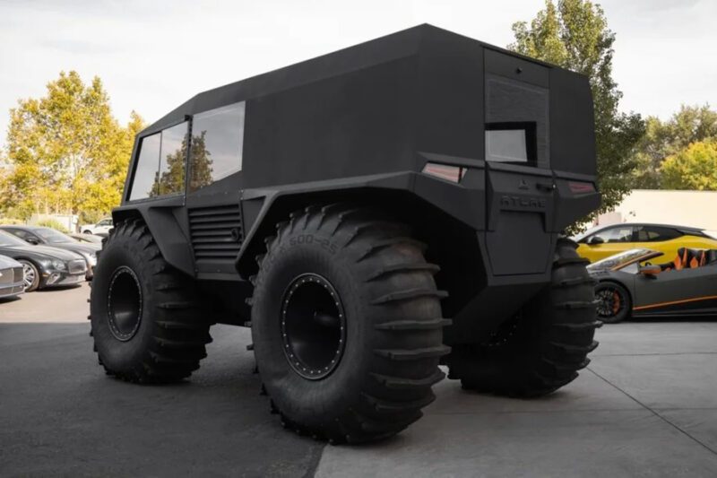 A large, angular black off-road vehicle with oversized tires is parked among other cars in a parking lot.