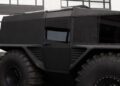 Black, heavily armored off-road vehicle with large tires parked in front of a building with glass windows.