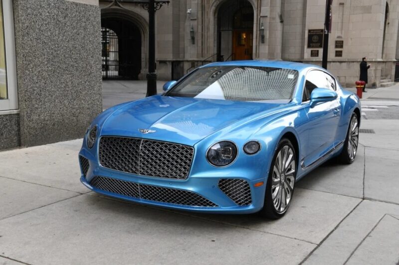 A shiny blue Bentley Continental GT, the epitome of a luxury tourer, is parked on a city street near a building entrance.