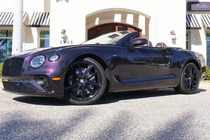 A dark purple Bentley Continental GTC convertible car is elegantly parked in front of a building with striped awnings.