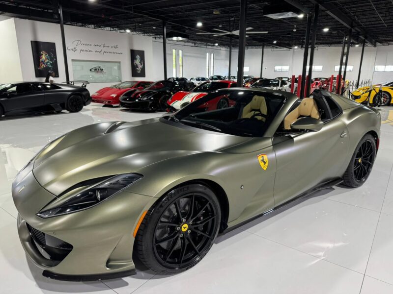 A matte olive green Ferrari, showcasing one of the unique Ferrari colors, sits on black wheels in a showroom filled with luxury and exotic cars.