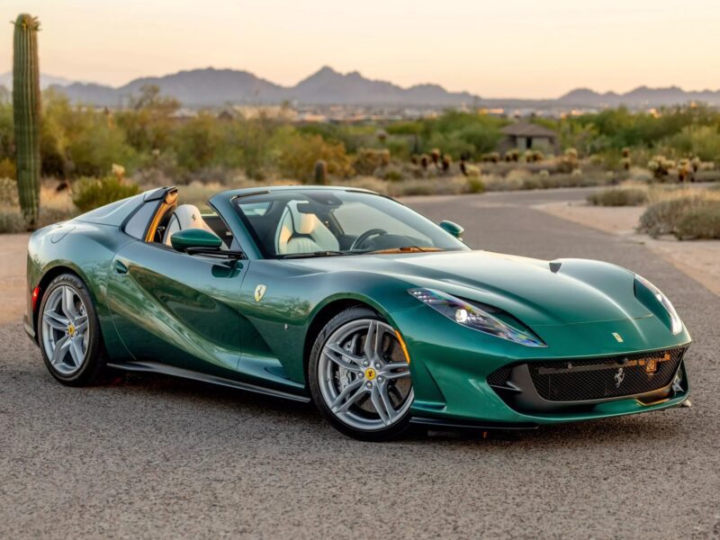 A green Ferrari convertible sports car, showcasing one of the Ferrari craziest colors, is parked on a paved road in a desert landscape with mountains in the background.
