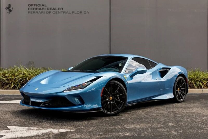 A unique Ferrari sports car in a striking blue hue is parked before a gray wall bearing the "Official Ferrari Dealer, Ferrari of Central Florida" signage, highlighting exclusive Ferrari colors.