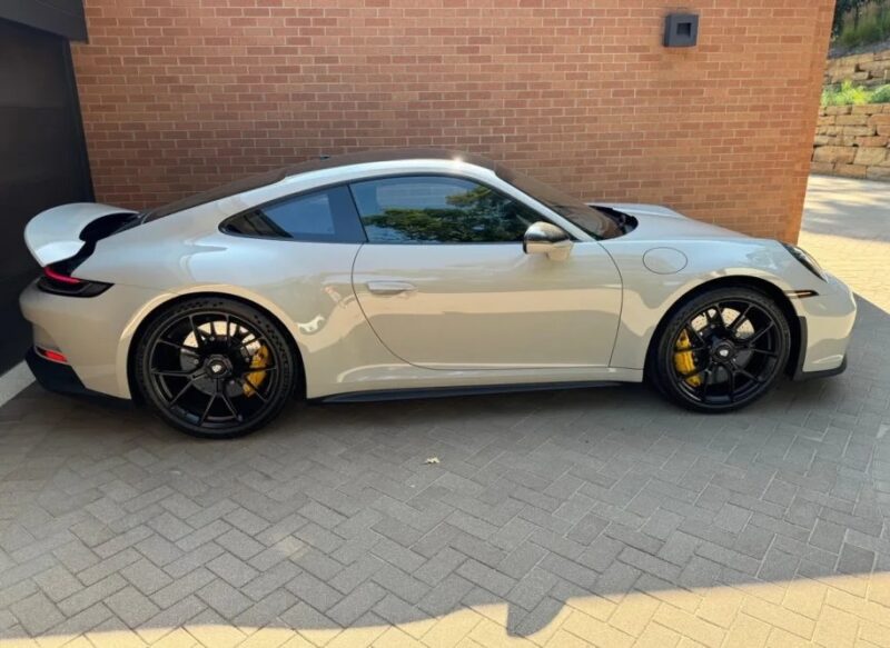 A gray sports car with black wheels and yellow brake calipers, renowned among the best manual cars, is parked on a brick driveway next to a brick wall.