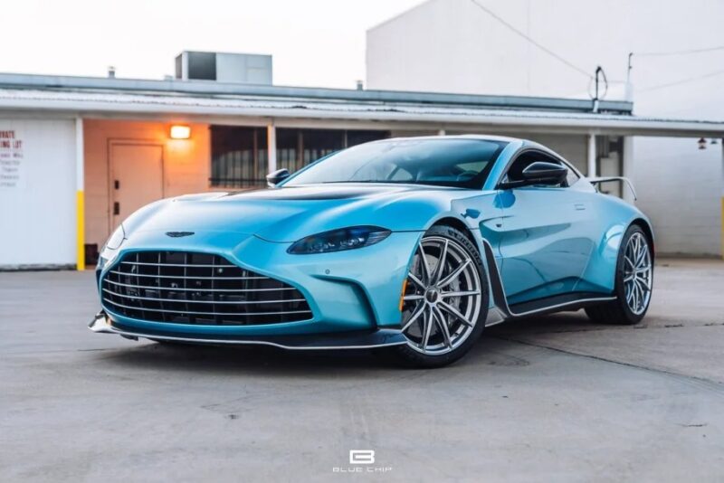 A sleek blue sports car, powered by a V12 engine, is parked in a lot with a modern building in the background.