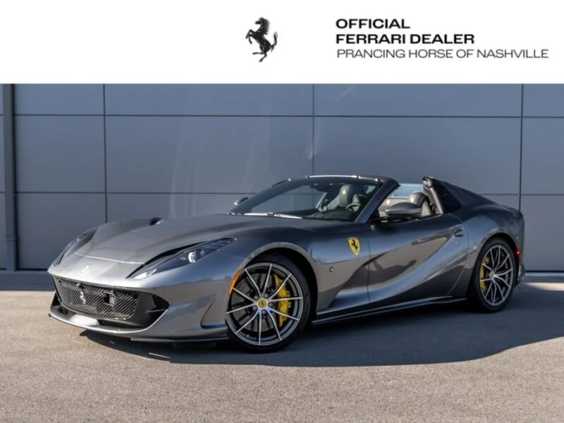 A sleek gray Ferrari convertible, featuring a powerful V12 engine, is parked on a paved surface, proudly displayed by an official Ferrari dealer.