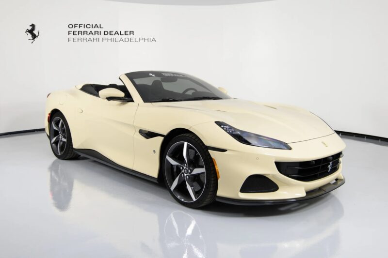 A cream-colored Ferrari Portofino convertible is displayed in a showroom with white walls, showcasing unique Ferrari colors and dealership branding on the wall behind—an unmistakable Ferrari for sale.