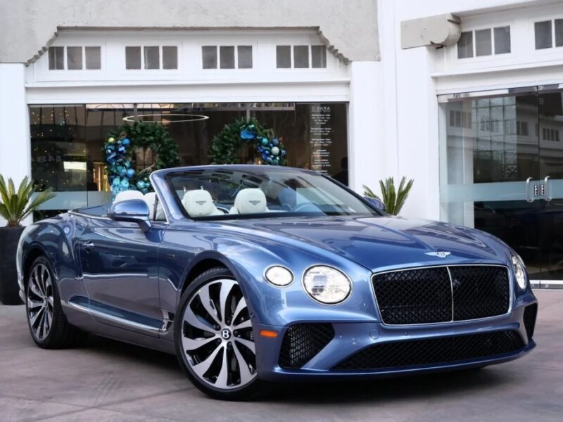 A luxurious Bentley convertible car is parked in front of a building with large windows and potted plants.