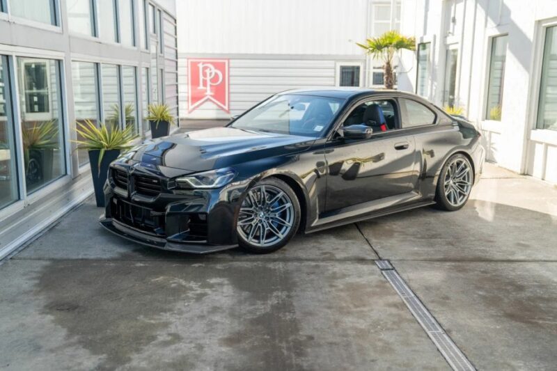 A sleek, black sports car with a manual transmission is parked on a concrete surface beside a modern building with large windows and plants.