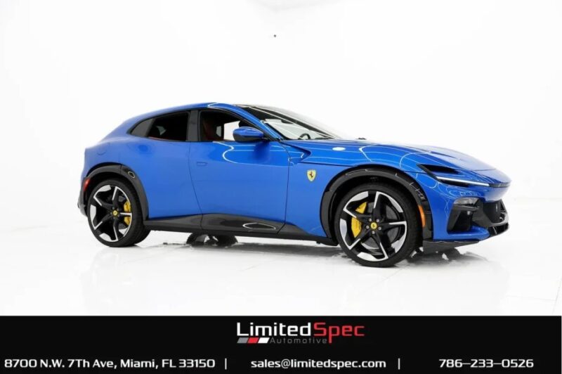 A blue sports car with a sleek design, featuring a powerful V12 engine, is displayed in the showroom. The car's doors are slightly open, showcasing yellow brake calipers and a logo on the side. This exotic car is now for sale.