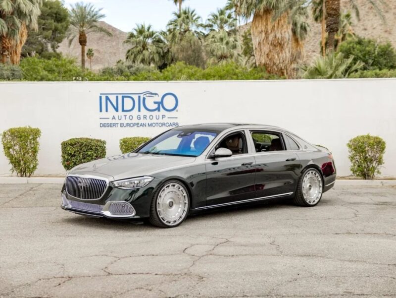 Sedan mewah Mercedes-Benz diparkir di depan dinding dengan "Indigo Auto Group Desert European Motorcars" tanda, dikelilingi oleh pohon -pohon subur dan pegunungan yang megah. Kendaraan mewah ini, tersedia untuk dijual, memancarkan keanggunan dan kecanggihan dengan latar belakang yang indah.