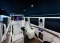 The interior of the 2024 Ultimate Class Jet Sprinter features white leather seats, ambient lighting, and a screen displaying an image, expertly crafted by First Class Customs for a refined travel experience.