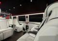 The 2024 Ultimate Class Jet Sprinter features a luxurious van interior with white leather seats, black accents, and a ceiling adorned with star-like lighting. A screen displays a person amid the opulent setting crafted by First Class Customs.