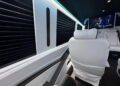 The interior of the 2024 Jet Sprinter exudes luxury with its white leather seats, ambient lighting, and a flat screen showcasing a scene from a show. Designed by First Class Customs, this vehicle redefines elegance on wheels.