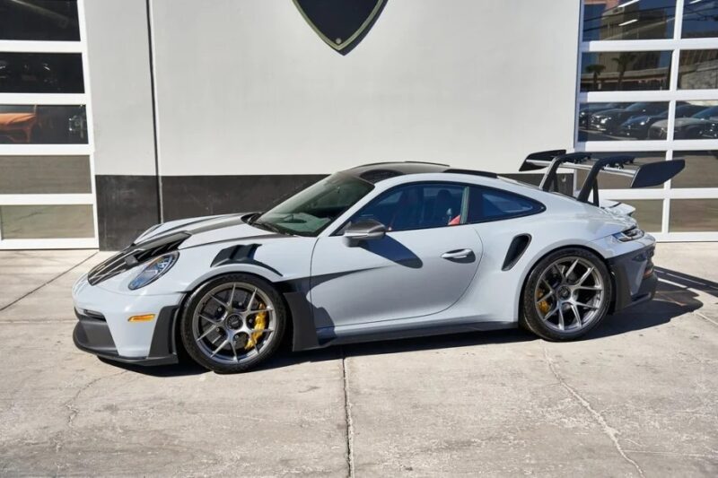 A gray Porsche 911 GT3 RS, renowned as Porsche's Ultimate Driver's Car, is parked in front of a building with large windows, its large rear spoiler accentuating its sleek design.