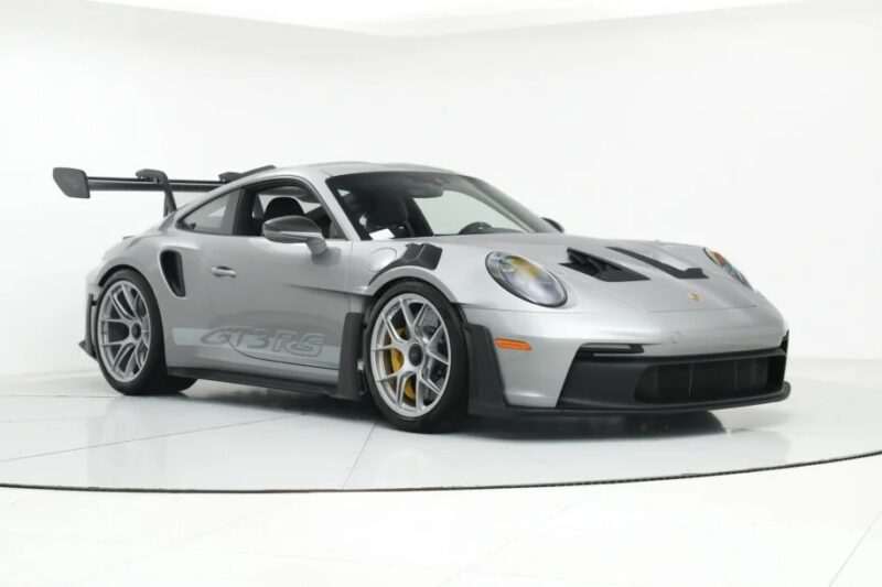 A silver Porsche 911 GT3 RS, the ultimate driver's car, with a large rear wing and aerodynamic features, is parked in a well-lit showroom.