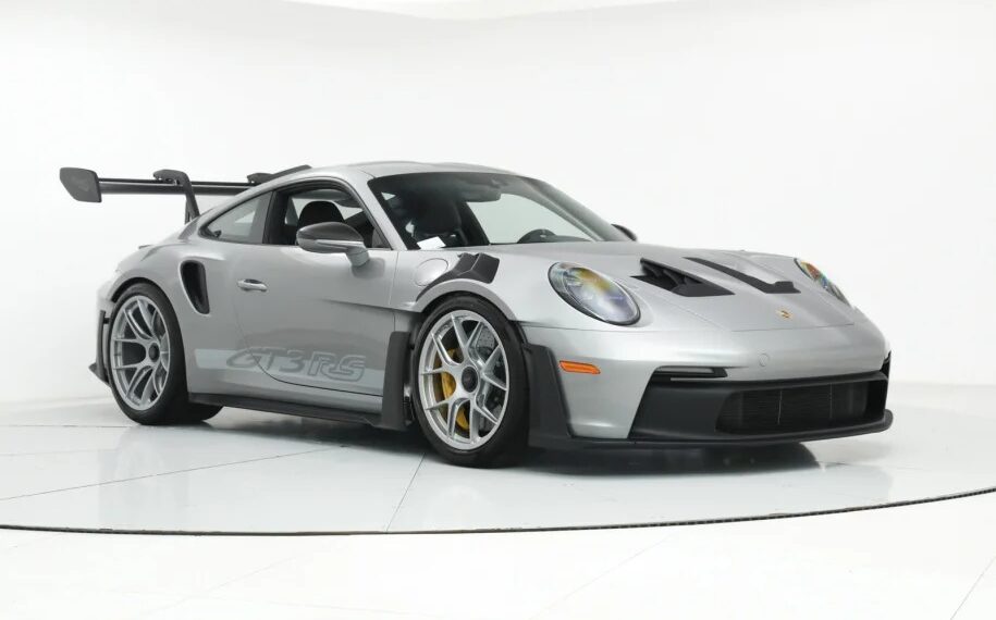 A silver Porsche 911 GT3 RS, the ultimate driver's car, with a large rear wing and aerodynamic features, is parked in a well-lit showroom.