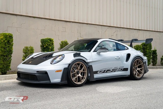 Porsche 911 GT3 RS, famously known as Porsche's Ultimate Driver's Car, gleams in silver with GT3 RS decals. The side view showcases brown wheels and a large rear spoiler, perfectly positioned near a beige building and vibrant green bushes.