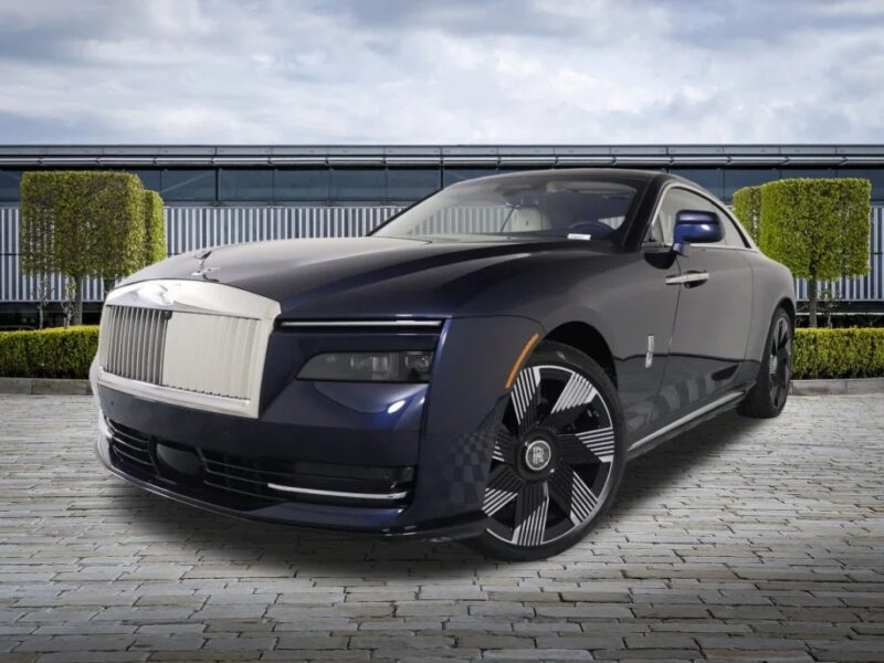 A Rolls-Royce Spectre in luxury blue and silver is elegantly parked on the stone pavement, framed by manicured hedges and a modern building. This ultra-luxe electric super coupe exudes sophistication and style in its serene setting.