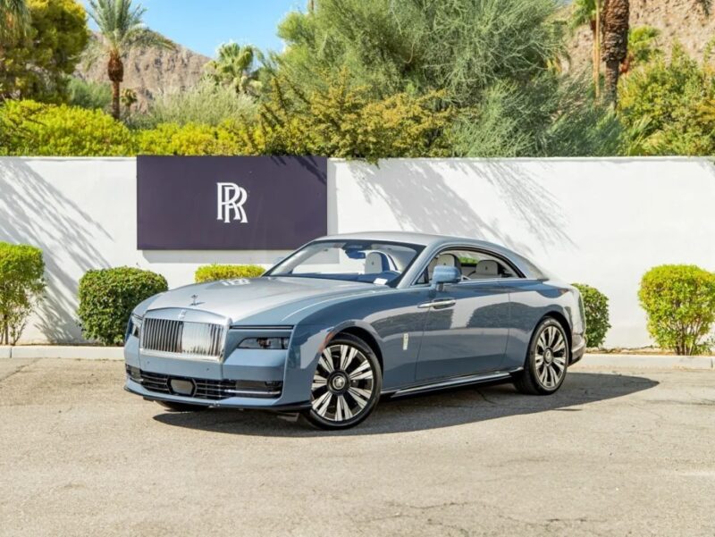 A blue Rolls-Royce Spectre, an ultra-luxe electric super coupe, is elegantly parked in front of a white wall with a black logo, surrounded by lush greenery.