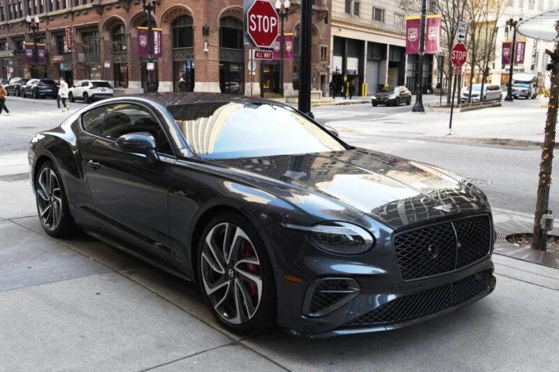 A sleek gray Bentley Continental GT is parked on a city street, near a stop sign and surrounded by tall buildings, epitomizing the essence of a luxury tourer.