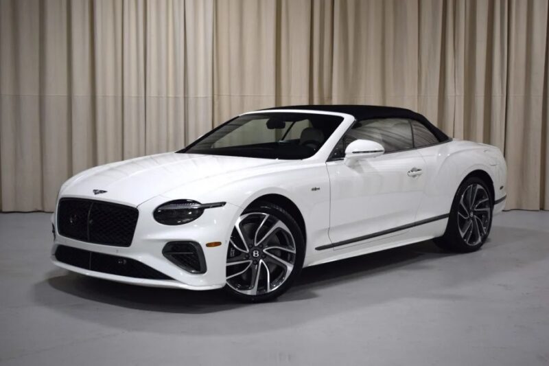 A luxurious Bentley Continental GTC convertible with a sleek black roof, elegantly parked indoors against a beige curtain backdrop.