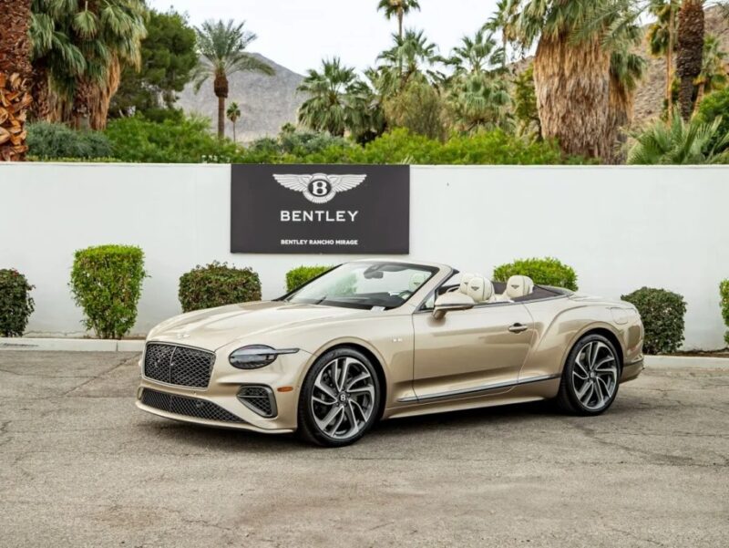 A beige Bentley Continental GTC convertible is elegantly parked in front of a Bentley dealership, surrounded by palm trees and hills, embodying the essence of a luxury tourer.