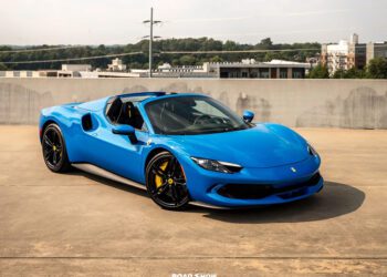 A blue Ferrari convertible sports car, showcasing one of the iconic Ferrari colors, is parked on a rooftop with city buildings and trees in the background.